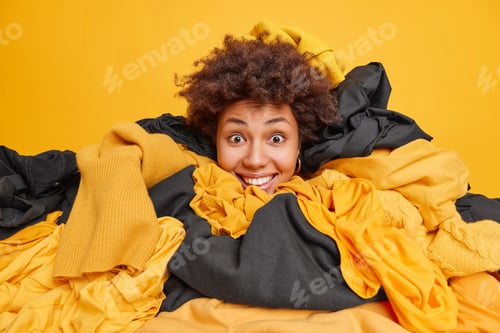 Preview: Positive Surprised Young Woman With Curly Hair Sticks Head In Big Pile Of Clothes Busy Cleaning Out