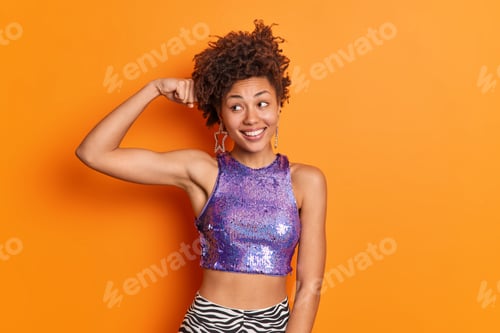 Preview: Fashionable Dark Skinned Afro American Woman With Curly Hair Raises Arm And Shows Muscles Wears