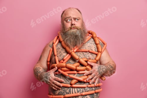 Preview: Puzzled Bearded Man Wrapped With Sausages Has Fat Tattooed Belly Excess Weight Isolated Over Pink