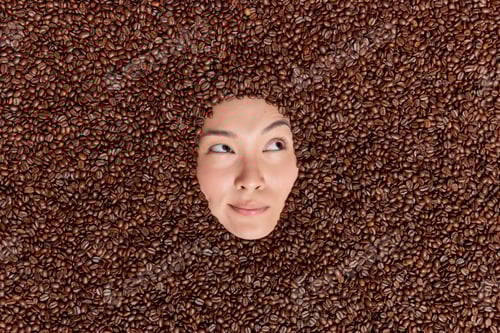 Preview: Pensive Female Model Drowned In Brown Coffee Beans Looks Thoughtfully Away Uses Roasted Seeds For