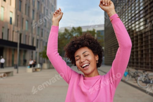 Preview: Carefree Positive Woman With Curly Hair Keeps Arms Raised Up Dances With Joy Walks At City Street