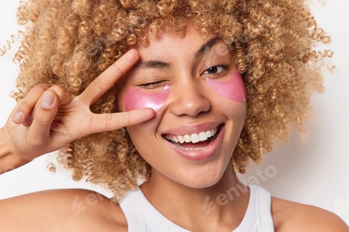 Preview: Headshot Of Positive Curly Haired Woman Smiles Broadly Winks Eyes Shows Peace Sign Undergoes Beauty