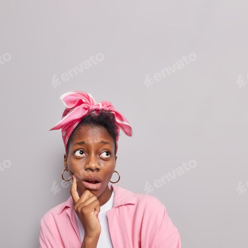 Preview: Vertical Shot Of Surprised Thoughtful Young Woman With Dark Skin Keeps Index Finger On Cheek
