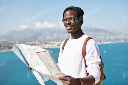 Preview: Happy Handsome Young Dark-Skinned Male Traveler Standing On Top Of Mountain With Paper Map Above