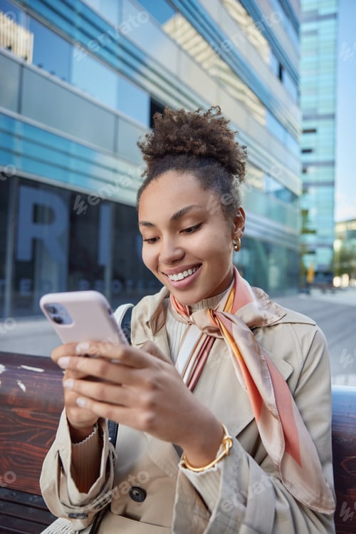 Preview: Happy Stylish Woman In Coat Sits On Wooden Bench Near Big Shopping Mall Uses Mobile Phone Chats