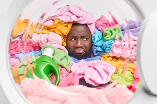 Preview: View From Washing Machine Of Upset Dark Skinned Bearded Man Drowned In Pile Of Multicolored Laundry