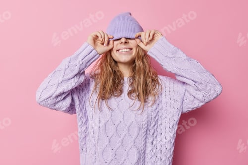 Preview: Happy Joyful Woman Covers Eyes With Hat Smiles Broadly Wears Purple Hat And Knitted Sweater Poses