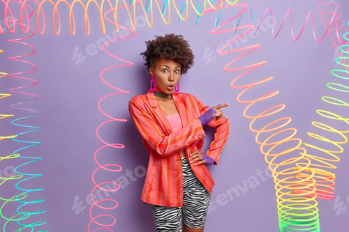 Preview: Impressed Dark Skinned Woman Points Aside Has Shocked Expression, Stands With Opened Mouth, Wears
