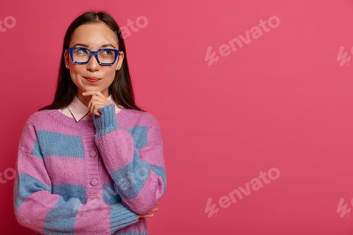 Preview: Thoughtful Brunette Female Student Wears Big Transparent Glasses, Looks To Right Side, Holds Chin