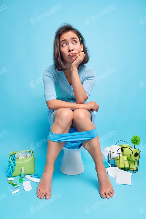 Preview: Vertical Shot Of Unhappy Thoughtful Woman Looks Away Sits On Toilet Bowl Suffers From Constipation