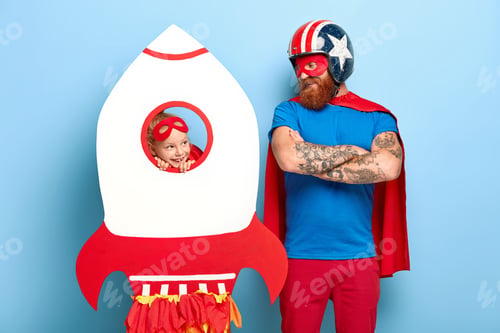 Preview: Horizontal Shot Of Satisfied Man Wears Headgear And Red Cape, Keeps Arms Folded, Looks At Playful