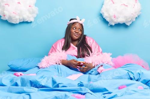 Preview: Pensive Dark Skinned Obese Woman Uses Modern Cellular For Chatting Online Wears Silk Dressing Gown