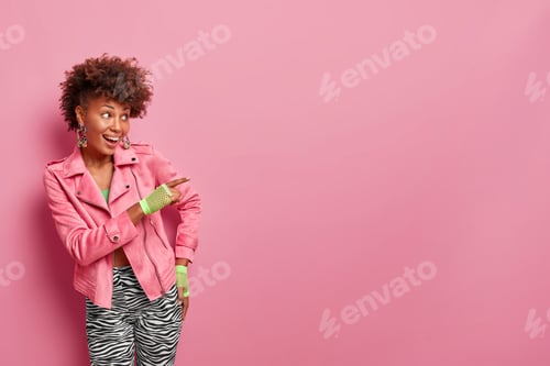 Preview: Positive Fashionable Curly Haired Woman Points Index Finger Away On Blank Space, Shows Promo