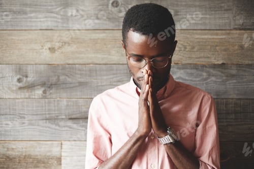 Preview: Young Meditating And Praying African American Man Wearing Pink Shirt And Glasses, Holding Hands In