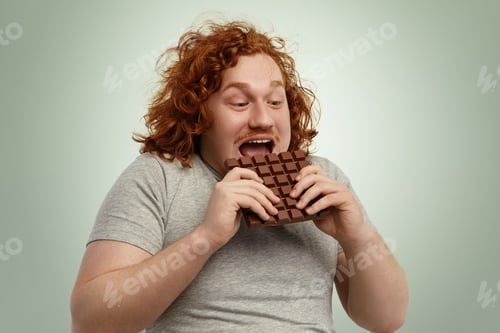 Preview: Happy Excited Young Chubby Red-Haired Male Opening Mouth Widely While Biting Bar Of Chocolate
