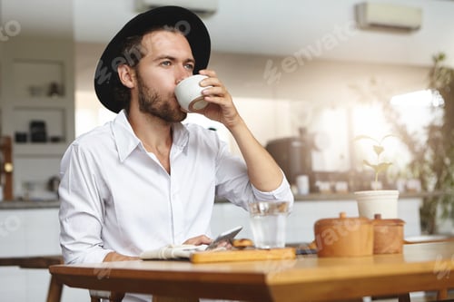 Preview: Fashionable Young Man With Beard Wearing Hat And White Shirt Having Hot Drink, Sitting At Table And