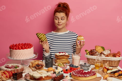 Preview: Pretty Redhead Woman Chooses Between Croissant And Cake, Sits At Table Overloaded With Desserts