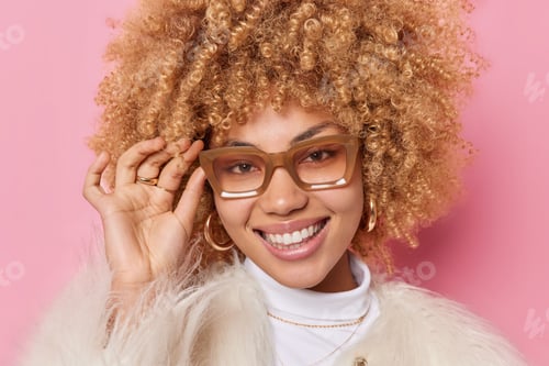 Preview: Portrait Of Cheerful Young Curly Woman Keeps Hand On Rim Of Spectacles Smiles Toothily Has Good