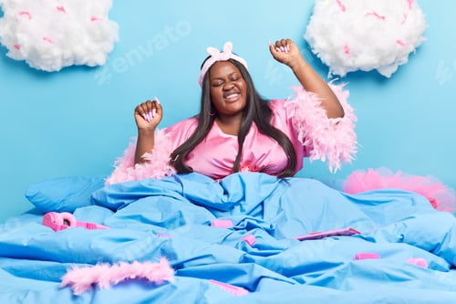 Preview: Happy Pleased Dark Skinned Plump Brunette Woman Has Fun In Bed Raises Arms And Feels Relaxed Smiles