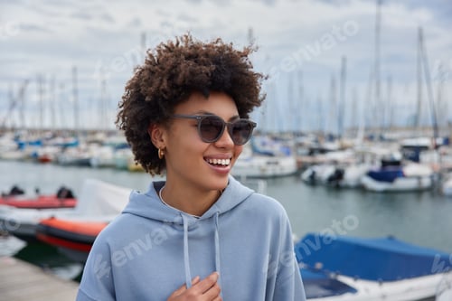 Preview: Happy Curly Haired Woman Wears Casual Hoodie Walks In Marina During Spare Time Enjoys Beautiful