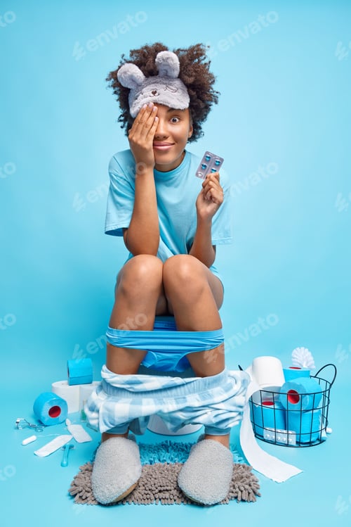 Preview: Indoor Shot Of Pleased Curly Haired Young Woman Covers Eye With Hand Holds Painkillers Poses On