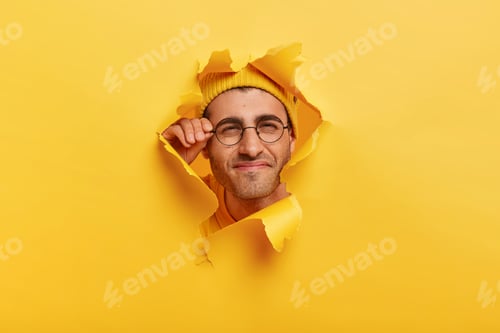 Preview: Male Face In Paper Torn Hole. Man Looks Curiously, Has Happy Gaze, Touches Frame Of Glasses, Poses