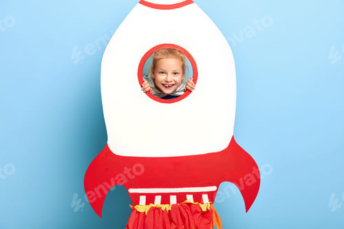 Preview: Lovely Kid Poses Behind Big Paper Rocket, Looks Through Round Hole, Has Toothy Smile And Happy