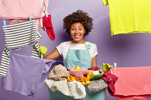 Preview: Smiling Dark Skinned Housekeeper Hangs Clean Clothes With Clothespegs On Washing Line, Happy To