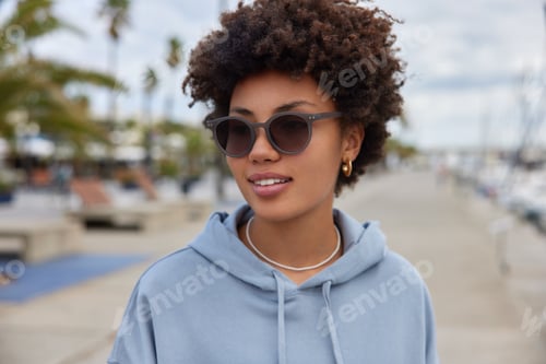 Preview: Outdoor Shot Of Pleased Woman With Curly Hair Wears Sunglasses And Hoodie Walks At Pier Harbor