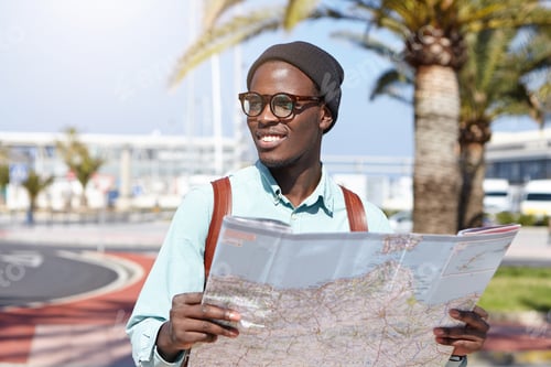 Preview: Happy Dark-Skinned Tourist In Trendy Hat And Shades Studying City Guide While Visiting New Town