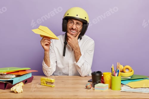 Preview: Discontent Office Worker Holds Yellow Paper Plane, Holds Chin, Wears Protective Helmet, Drinks