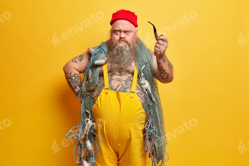 Preview: Angry Irritated Bearded Boatswain Looks With Sullen Expression Directly At You Holds Smoking Pipe