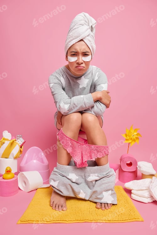 Preview: Indoor Shot Of Unhappy Young Woman Dressed In Pajama Suffers From Constipation Poses On Toilet Bowl