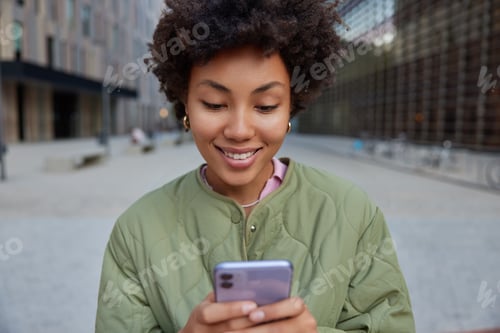 Preview: Pretty Smiling Hipster Girl With Afro Hair Holds Digital Smartphone Surfs Social Media Wears Jacket