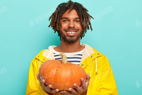 Preview: Cheerful Dark Skinned Man In Yellow Raincoat Boasts His Autumn Crop, Holds Pumkin, Smiles