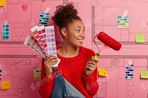 Preview: Pleased Female Painter With Curly Combed Hair Looks Aside, Notices Something Nice, Stands With