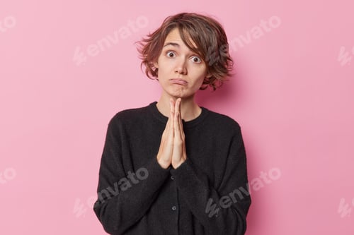 Preview: Coy Beautiful Woman With Short Hair Keeps Hands In Pray Beg Gesture Says Please Wears Casual Black