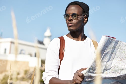 Preview: Outdoor Shot Of Attractive Young Black Male Tourist In Trendy Sunglasses And Hat Holding Paper Map