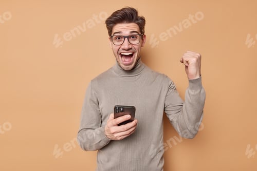 Preview: Emotional Handsome Man Clenches Fist In Victory And Cheer Gesture Reads Awesome News Via Smartphone