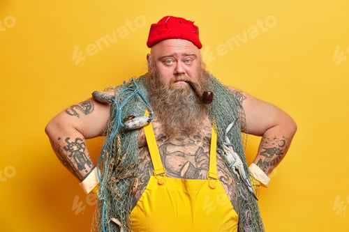 Preview: Serious Plump Tattooed Man Sailor Stands In Self Confident Pose Keeps Hands On Waist Smokes Pipe