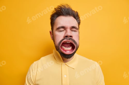 Preview: Studio Shot Of Bearded Tired Caucasian Man Closes Eyes Keeps Mouth Widely Opened While Yawning Has