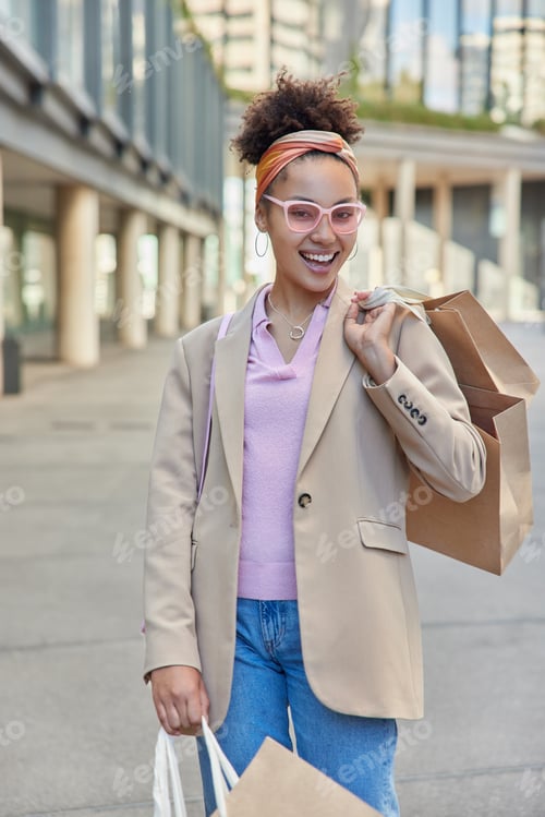 Preview: Happy Young Woman Enjoys Holiday Discounts In Shopping Mall Walks At Street With Paper Bags Wears
