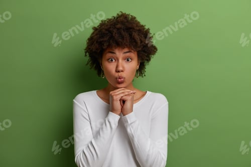 Preview: Studio Shot Of Lovely Dark Skinned Woman Folds Lips In Air Kiss And Keeps Hands Under Chin, Wears