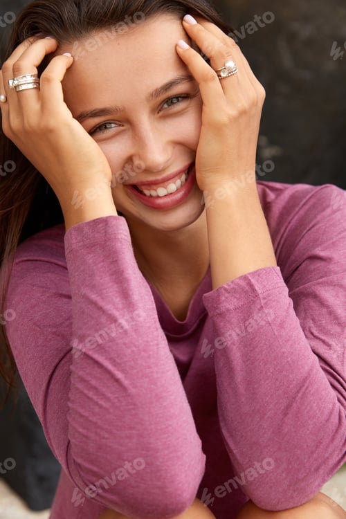 Preview: Joyful Young Model With Pleasant Smile, Keeps Hands On Face, Being In High Spirit, Looks Positively