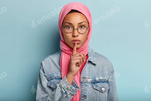 Preview: Headshot Of Serious Female In Pink Scarf, Denim Coat, Keeps Fore Finger Over Lips, Asks Not Make