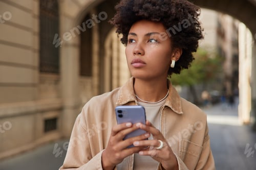 Preview: Outdoor Shot Of Thoughtful Young Woman Surfs Social Media While Strolling In Urban Street Wears