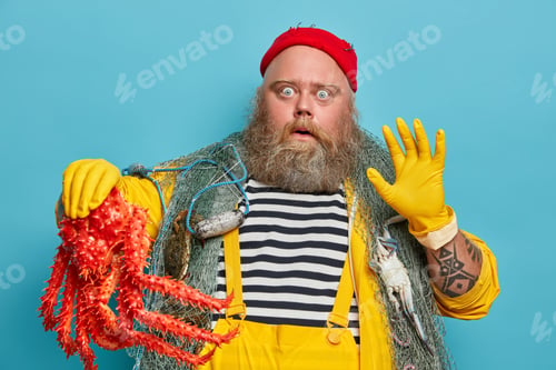 Preview: Plump Bearded Fisherman Stares With Bugged Eyes, Being Scared, Raises Hand, Wears Yellow Rubber