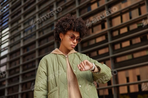 Preview: Photo Of Serious Curly Woman Hurries For Work Or Meeting Looks Attentively At Wrist Watch Checks