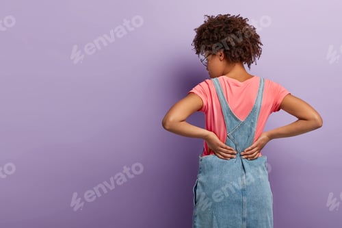 Preview: Horizontal Shot Of Dark Skinned Curly Woman Stands Backwards At Camera, Keeps Hands On Waist, Wears