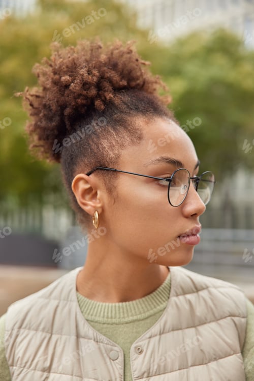 Preview: Profile Shot Of Curly Haired Clever Woman Wears Round Spectacles And Vest Focused Away Into